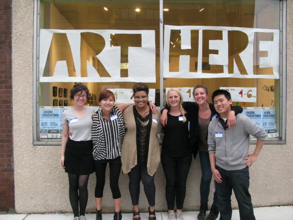 Irrigate interns standing abreast in front of gallery window that says -Art Here.
