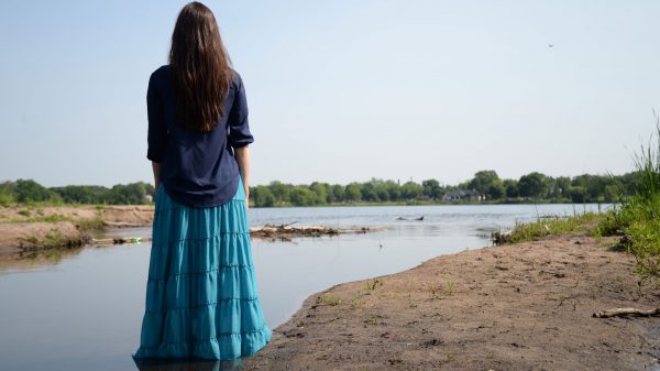 A dancer in a dark blue top and light blue skirt from Wisdom Dances looks out over a river