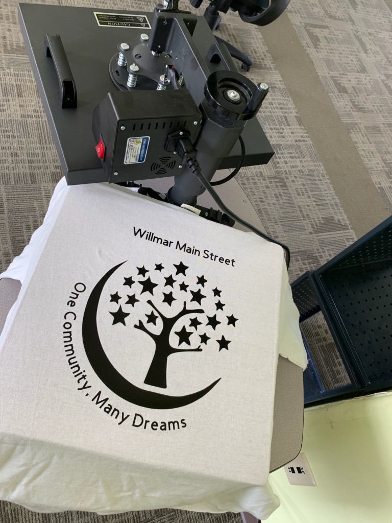 A t-shirt being printed with the words "Willmar Main Street, One Community, Many Dreams" and a design of a tree with stars for leaves surrounded by a crescent moon.