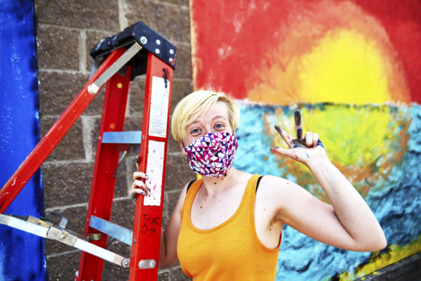 A woman with a mask on, holding a ladder, with paint-stained hands raised in front of a mural