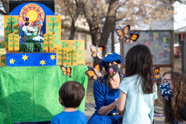 A performer in a blue mask with monarch butterfly puppets in front of a colorful set