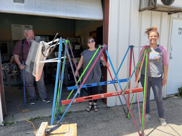 Three artists stand outside a building, next to an abstract, colorful metal sculptural shape