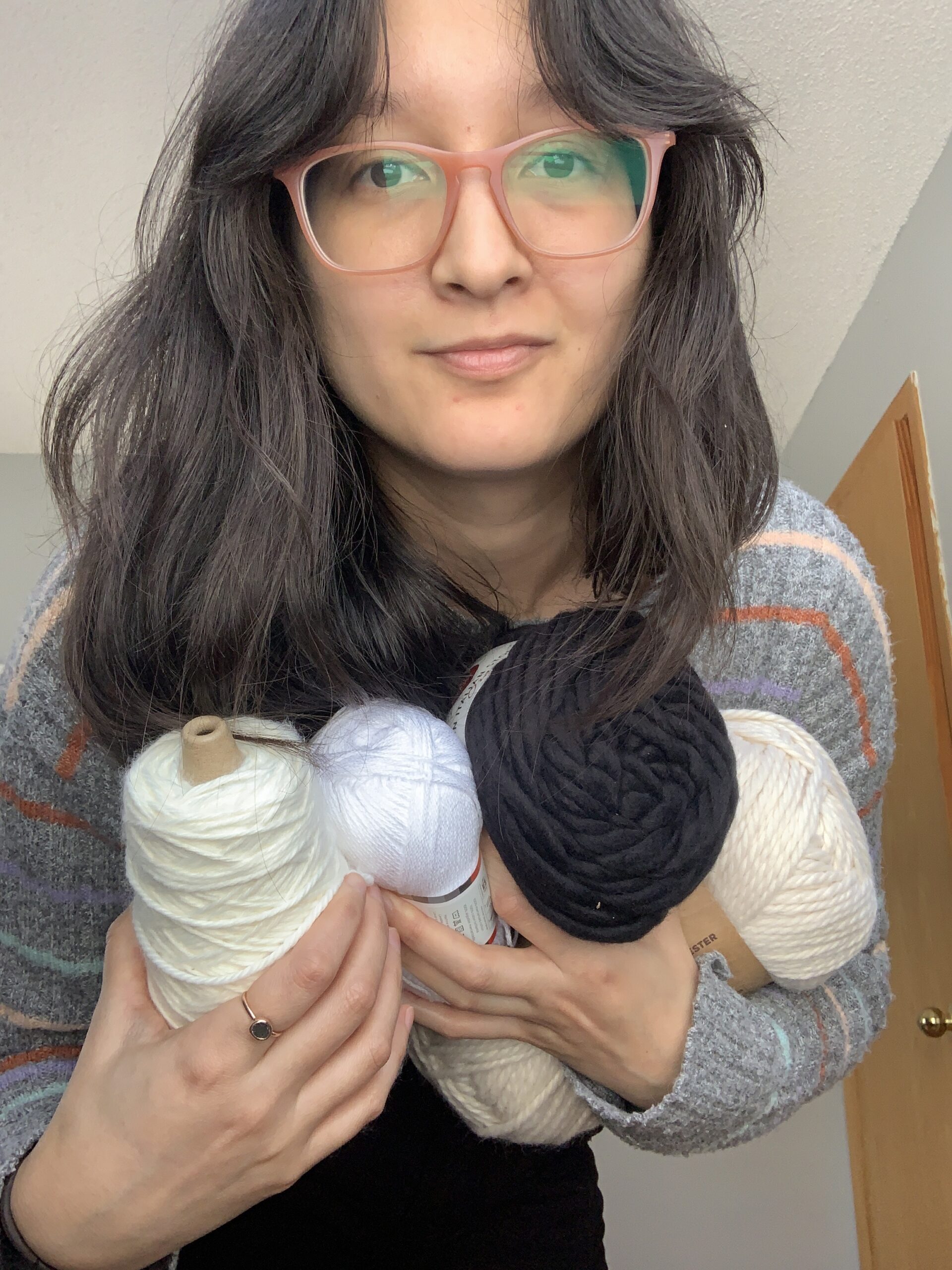 Nicole is a tan skinned Vietnamese American with shoulder length wavy hair with arms full of yarn skeins. They are wearing a striped sweater with pink square glasses and looking directly at the camera with a soft smile.