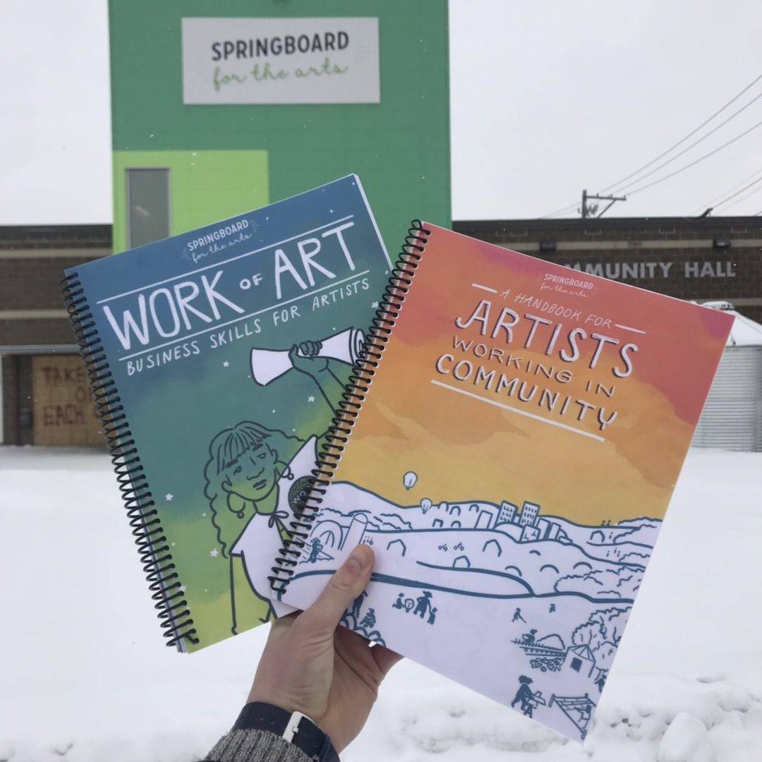 A photo of a hand holding up Springboard Books, Work of Art: Business Skills for Artists, and the Handbook for Artists Working in Community